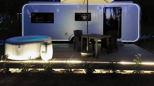 Una autocaravana moderna con bañera de hidromasaje, mesa, sillas y una sombrilla.