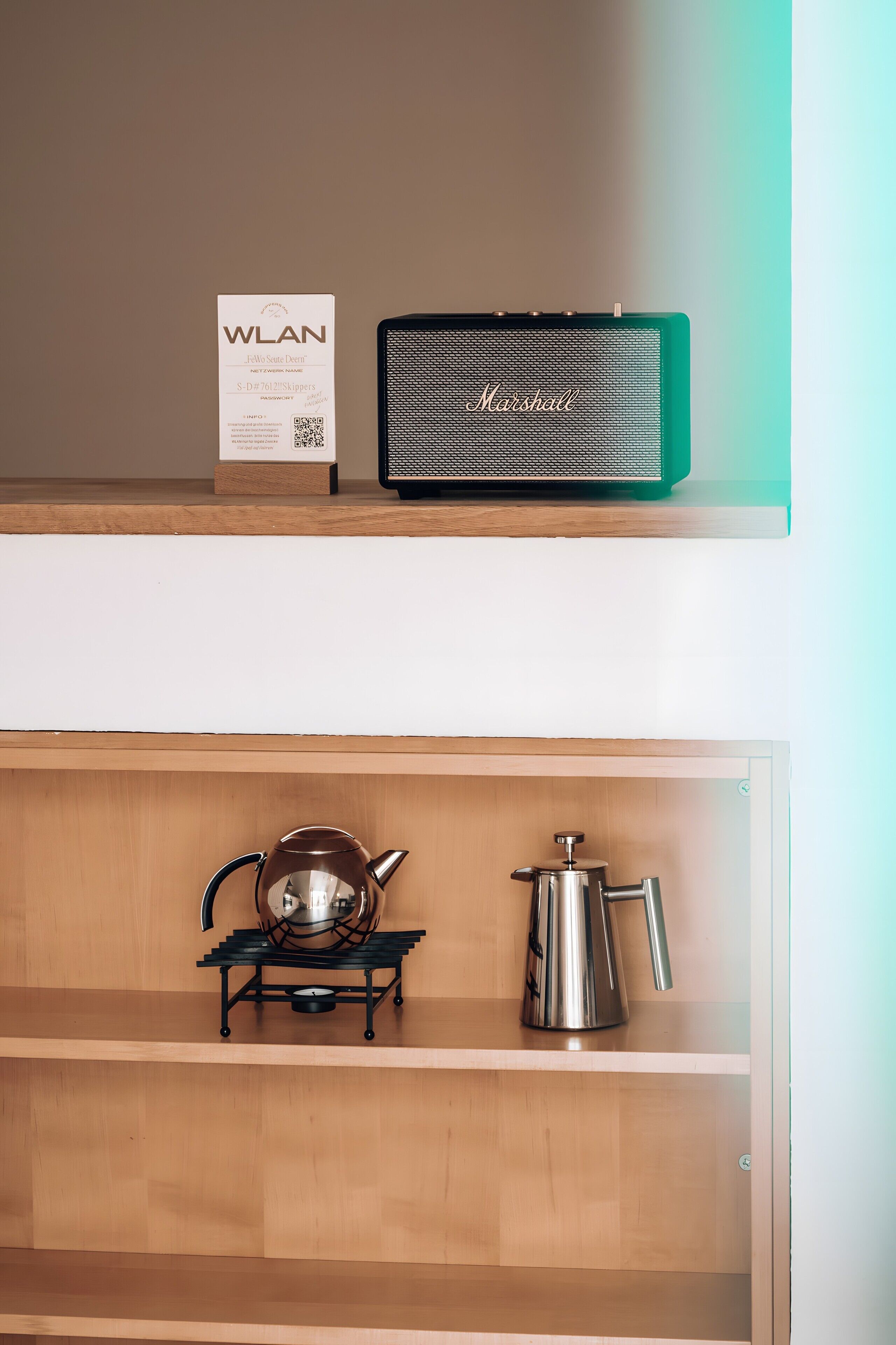 A kitchen area with a coffee maker, a kettle, a box labeled 'WLAN,' and a green speaker on a shelf.