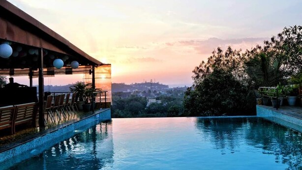 Una piscina infinita en la azotea con vista a la ciudad al atardecer, mobiliario de terraza de madera y plantas en macetas.