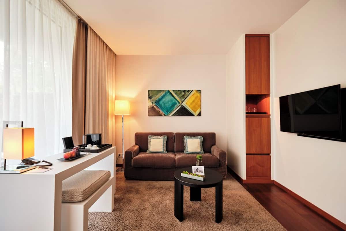 A modern hotel room with a sofa, a coffee table, a desk with a lamp, a TV, and a painting on the wall.