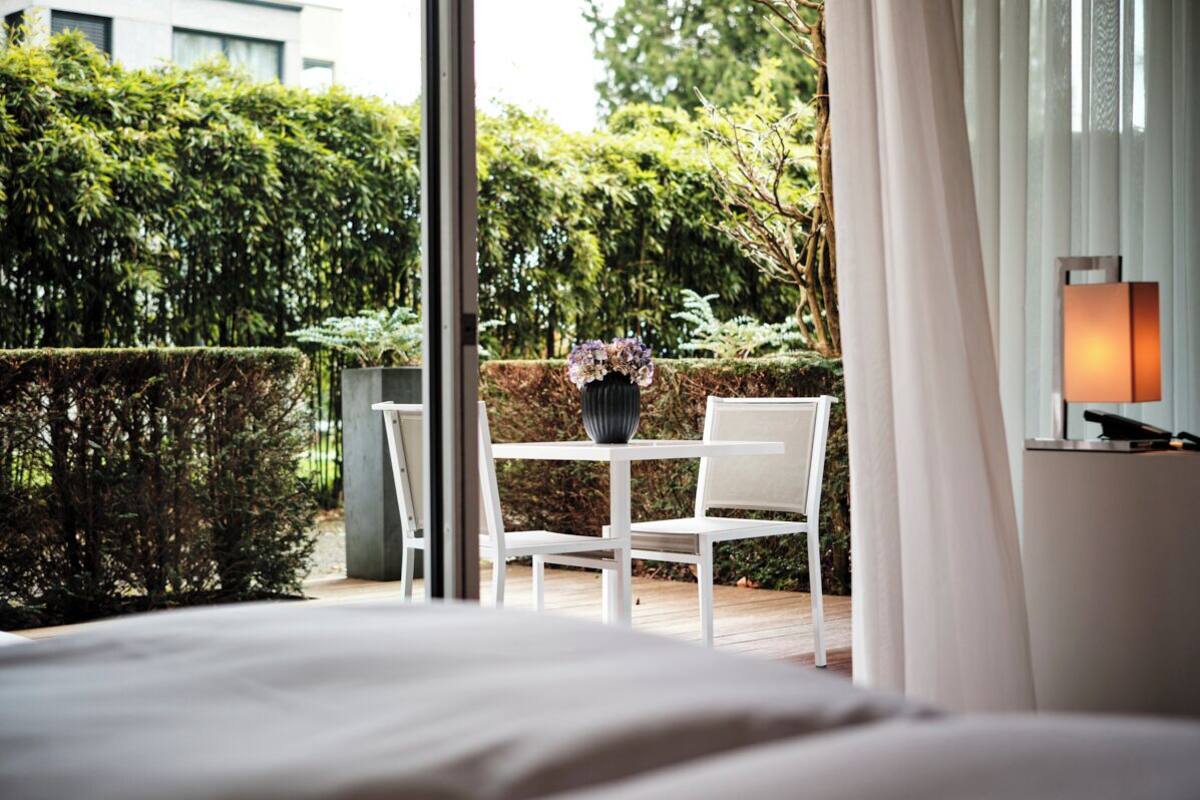 A hotel room with a bed, a table with a vase of flowers, and a view of a garden through a window.