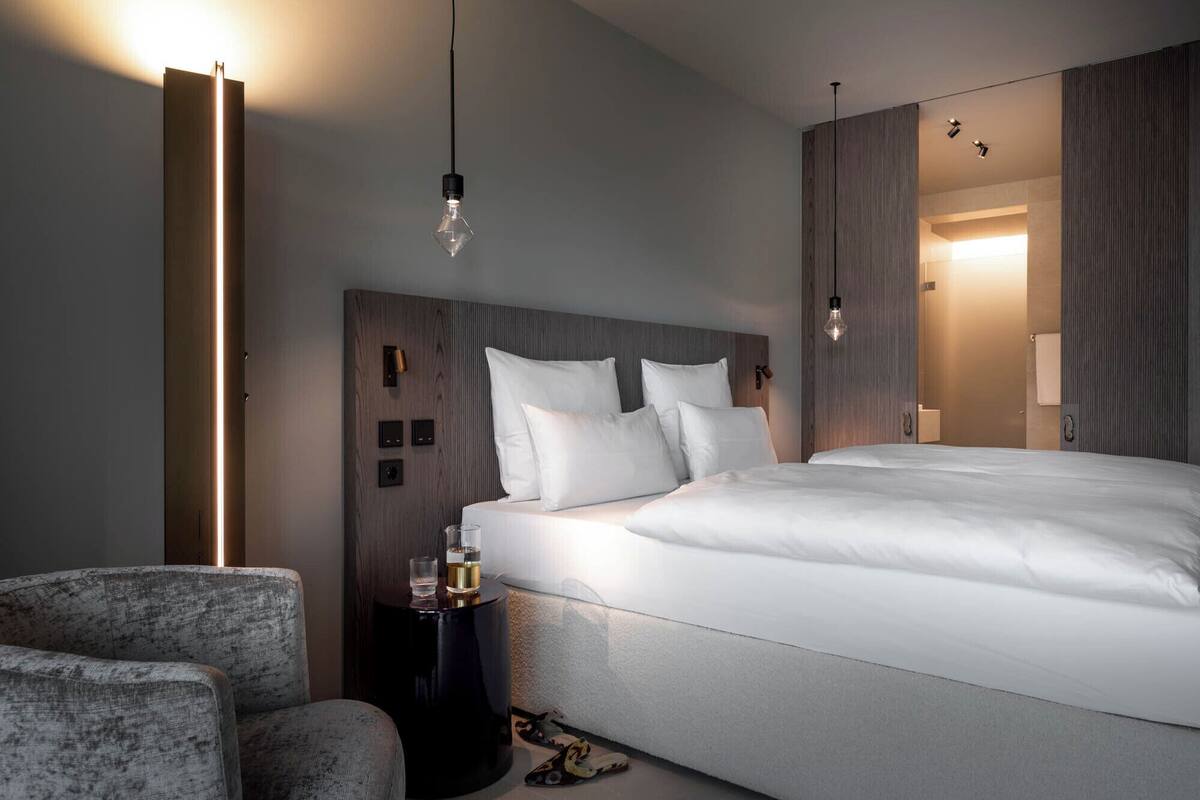 A modern hotel room with a large bed, a side table, a chair, and a wall-mounted light fixture.