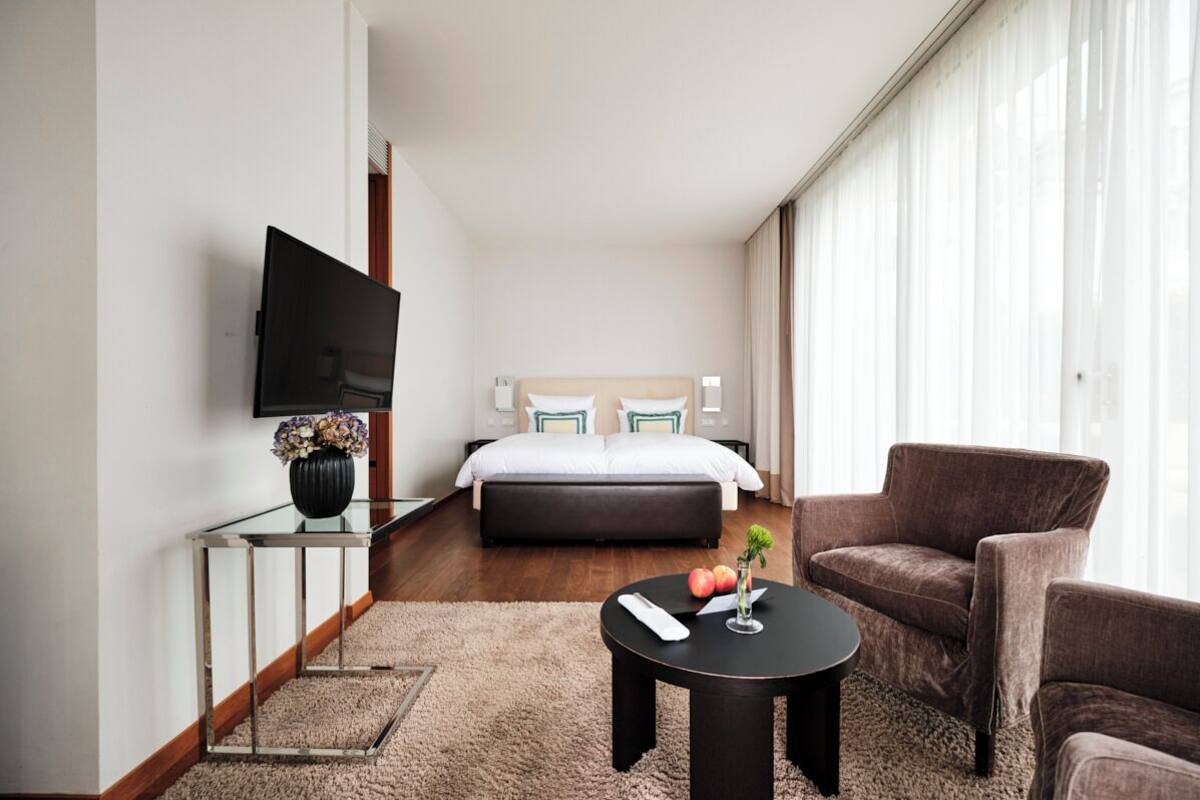 A modern hotel room with a large bed, a TV, a small table with a vase, and a chair.