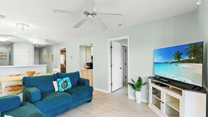 Un salon avec un canapé de couleur turquoise, un ventilateur de plafond, un téléviseur à écran plat posé sur un meuble en bois, et une cuisine en arrière-plan.