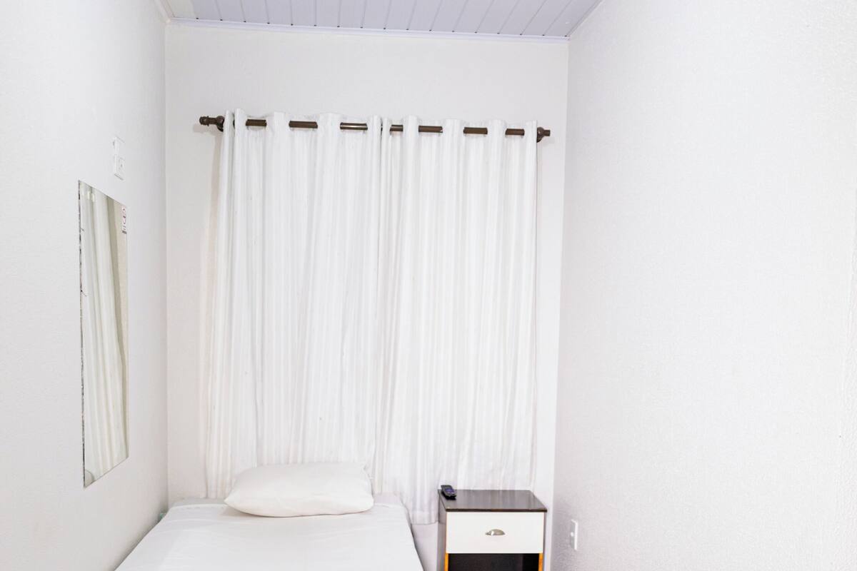 A small, clean room with a single bed, a nightstand, a mirror, and a window with white curtains.