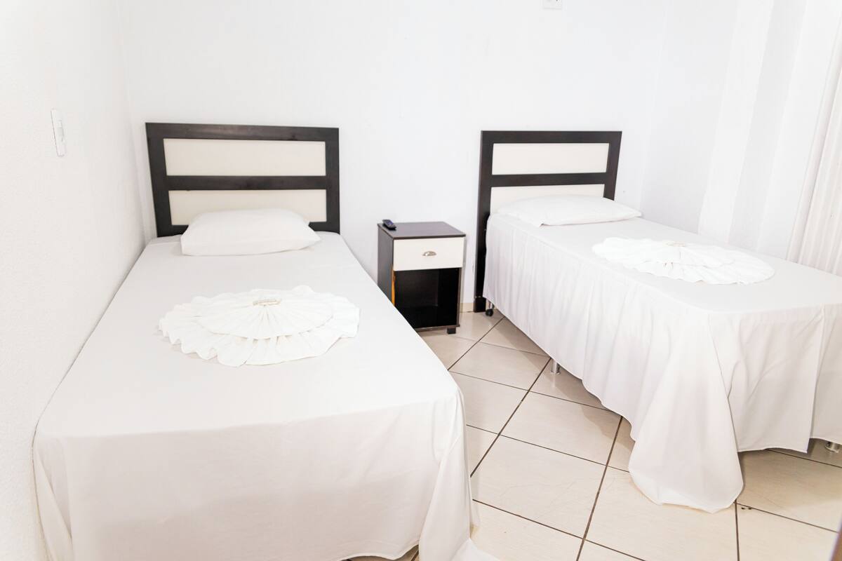 A hotel room with two single beds, each with a white bedspread and a decorative pillow. There is a small bedside table with a lamp and a drawer.