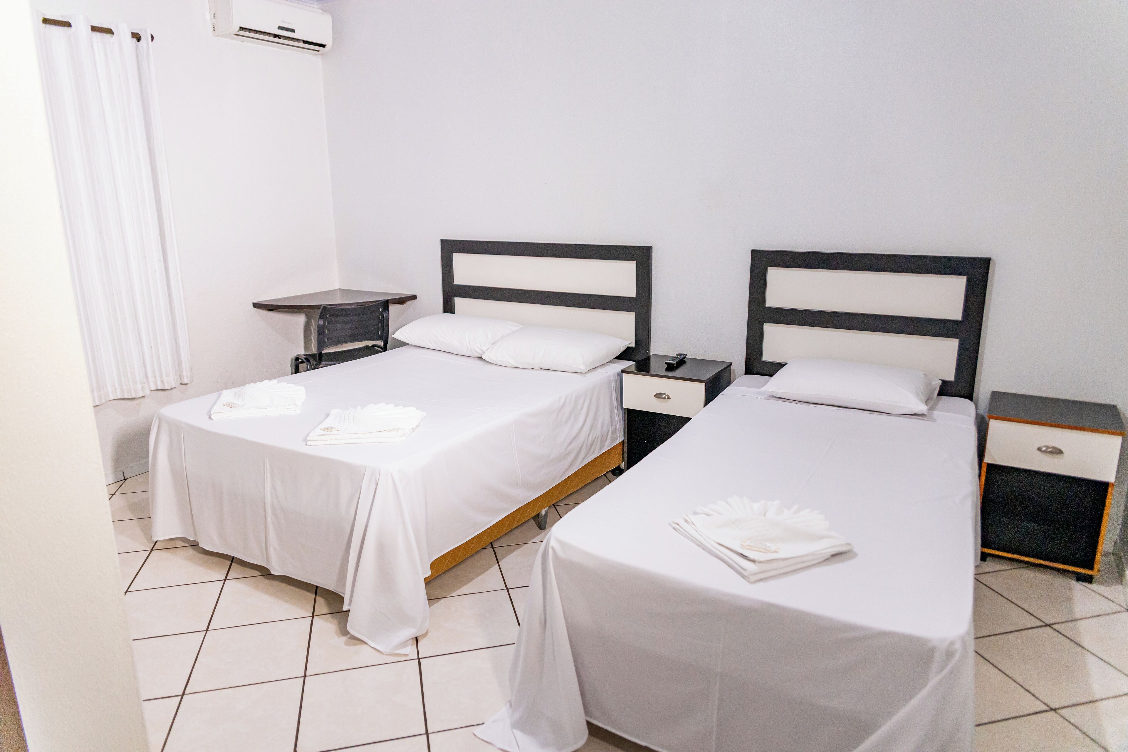 Habitación de hotel con dos camas, cada una con cubrecama y almohadas blancas, una mesita de noche con lámpara y una pequeña cómoda.