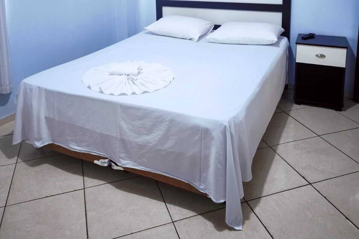 A neatly made bed with white linens and pillows, a bedside table with a drawer, and a tiled floor.