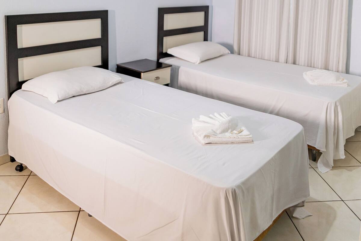 A hotel room with two single beds, a nightstand, a small table, a ceiling fan, and a window with curtains.