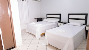 A hotel room with two single beds, a bedside table with a lamp, a small desk, and a window with curtains. - Hotel Village (Pato Branco)