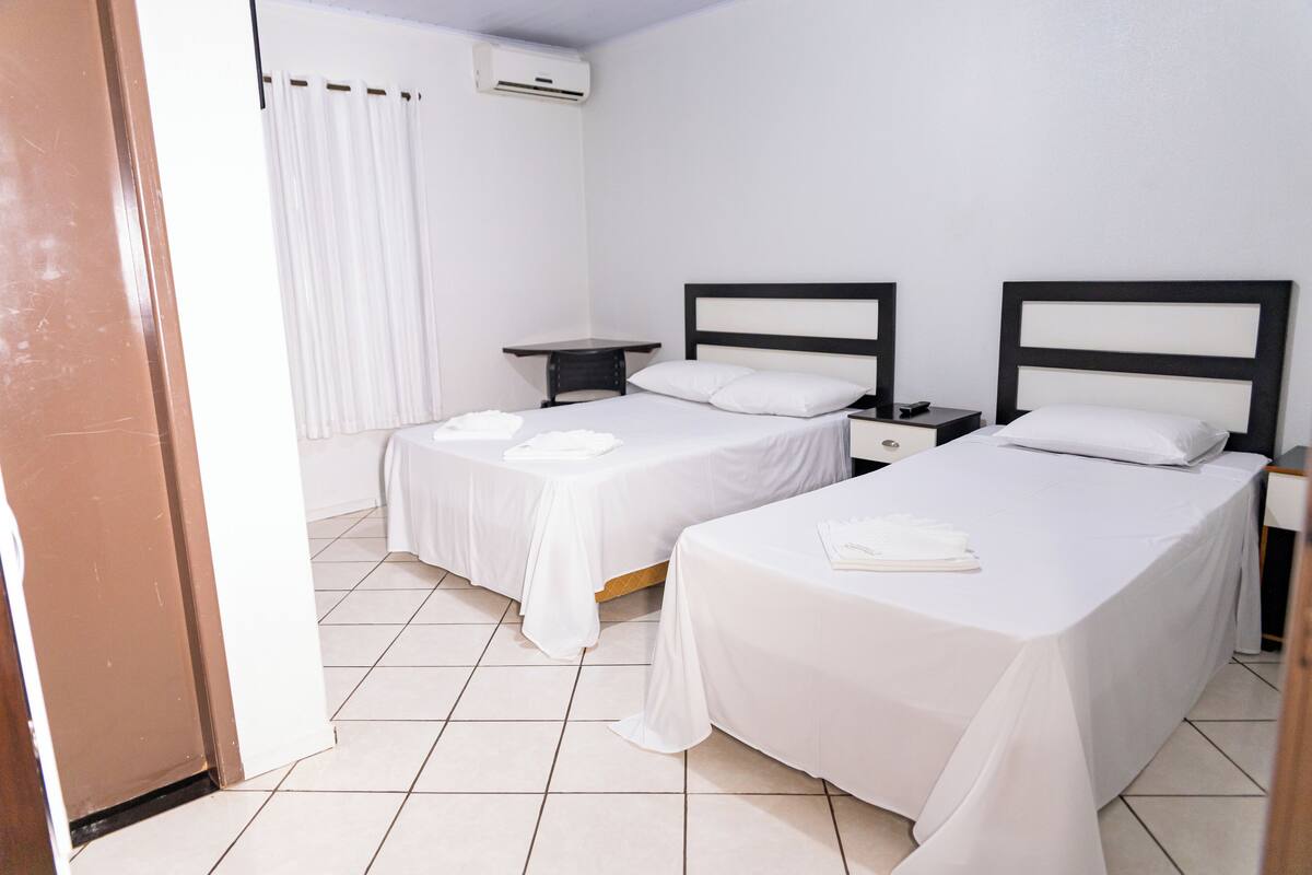 A hotel room with two single beds, a bedside table with a lamp, a small desk, and a window with curtains.