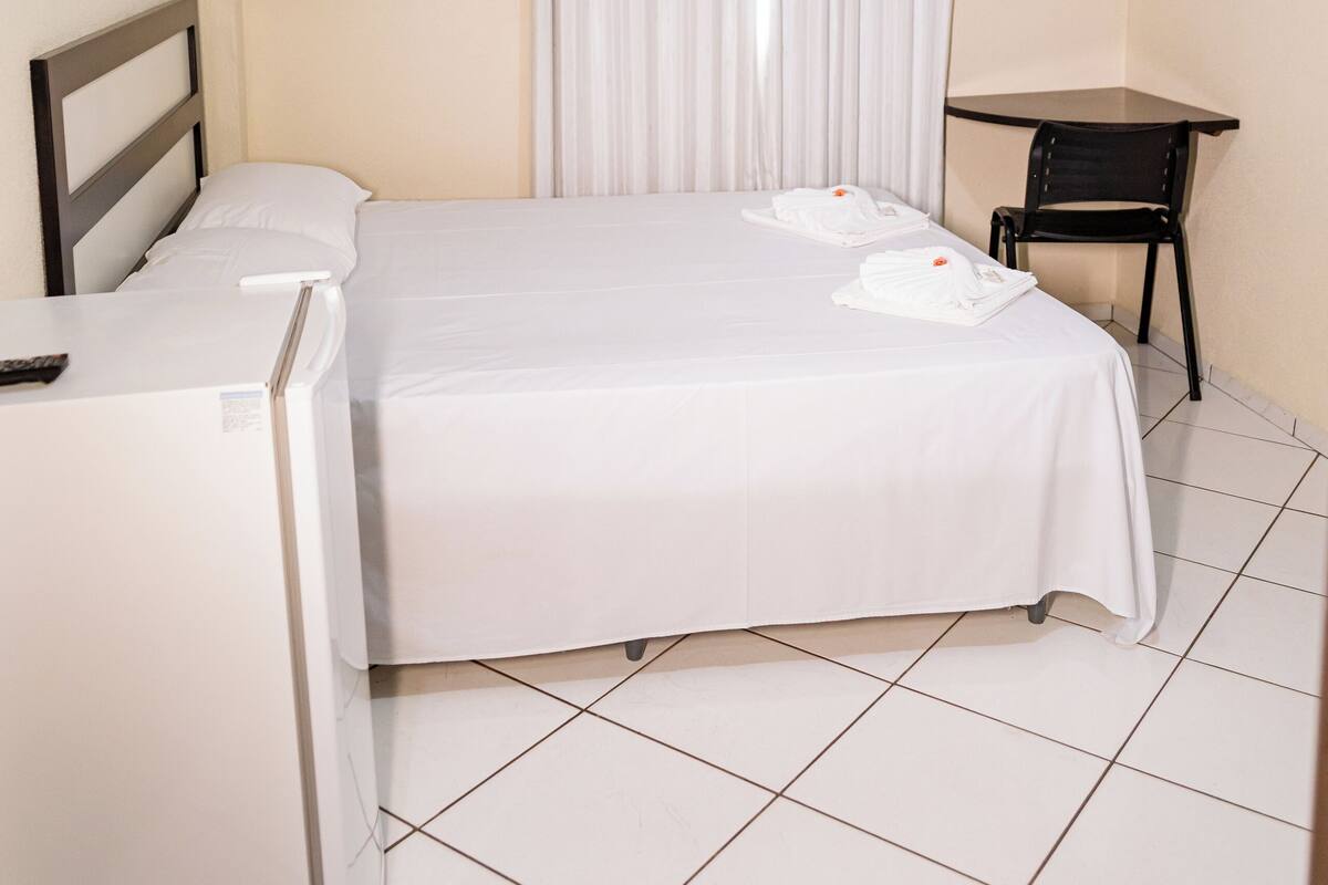 A hotel room with a bed, a desk, a chair, a TV, and an air conditioning unit.