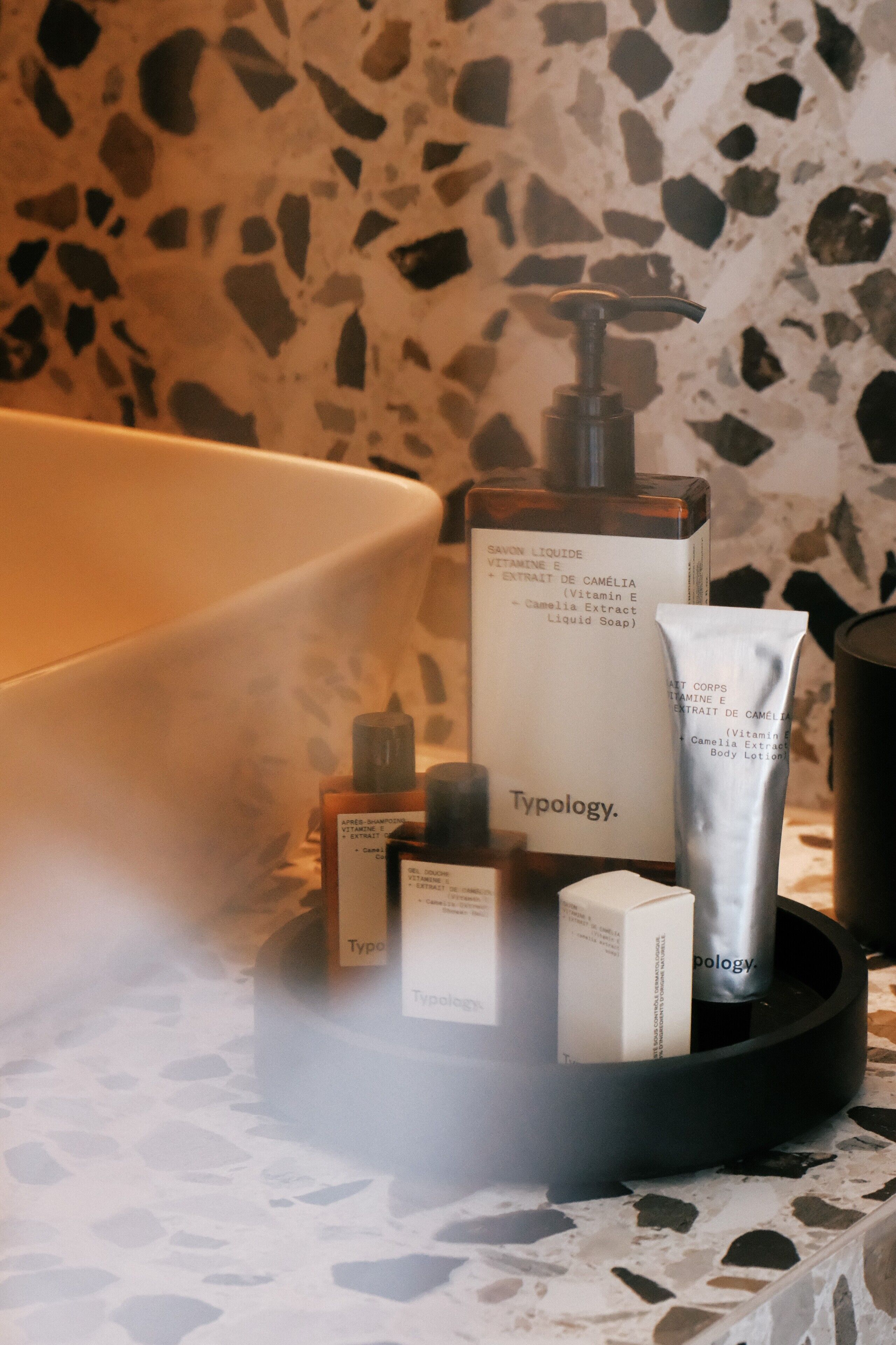 Un meuble-lavabo de salle de bain avec des articles de toilette, notamment un flacon de lotion pour le corps de la marque Typology, un tube de crème et un flacon-pompe, le tout placé devant un mur recouvert de carreaux à motifs.