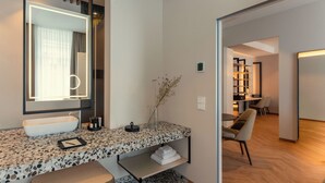 A modern bathroom with a large mirror, a white sink, and a vanity with a terrazzo countertop. There is a bench in the corner and a small shelf with towels. - LOVIS Suites Vienna - NEW OPENING (Vienna)