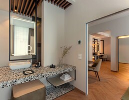 A modern bathroom with a large mirror, a white sink, and a vanity with a terrazzo countertop. There is a bench in the corner and a small shelf with towels.