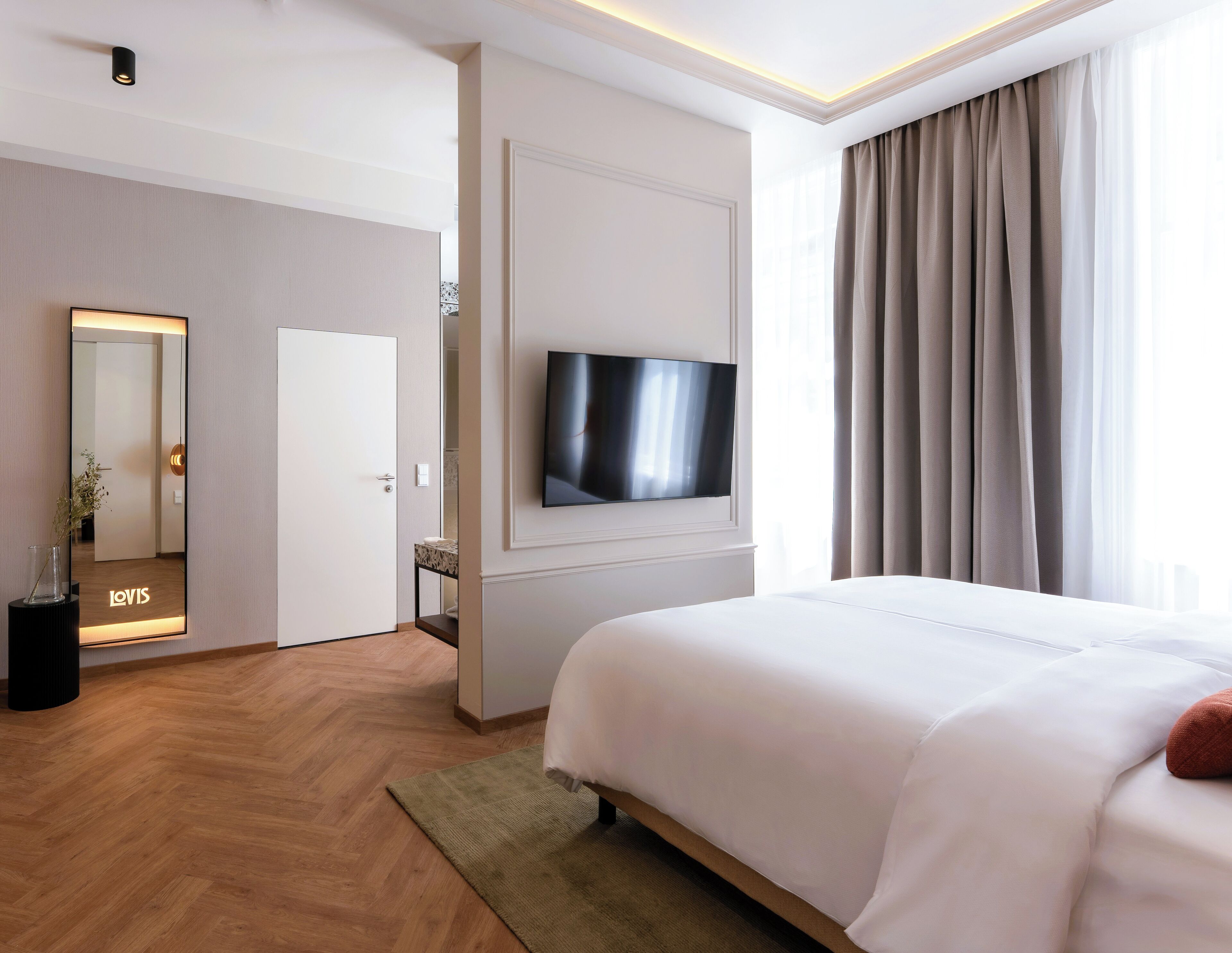 A hotel room with a bed, a TV mounted on the wall, a mirror, and a door leading to another room.