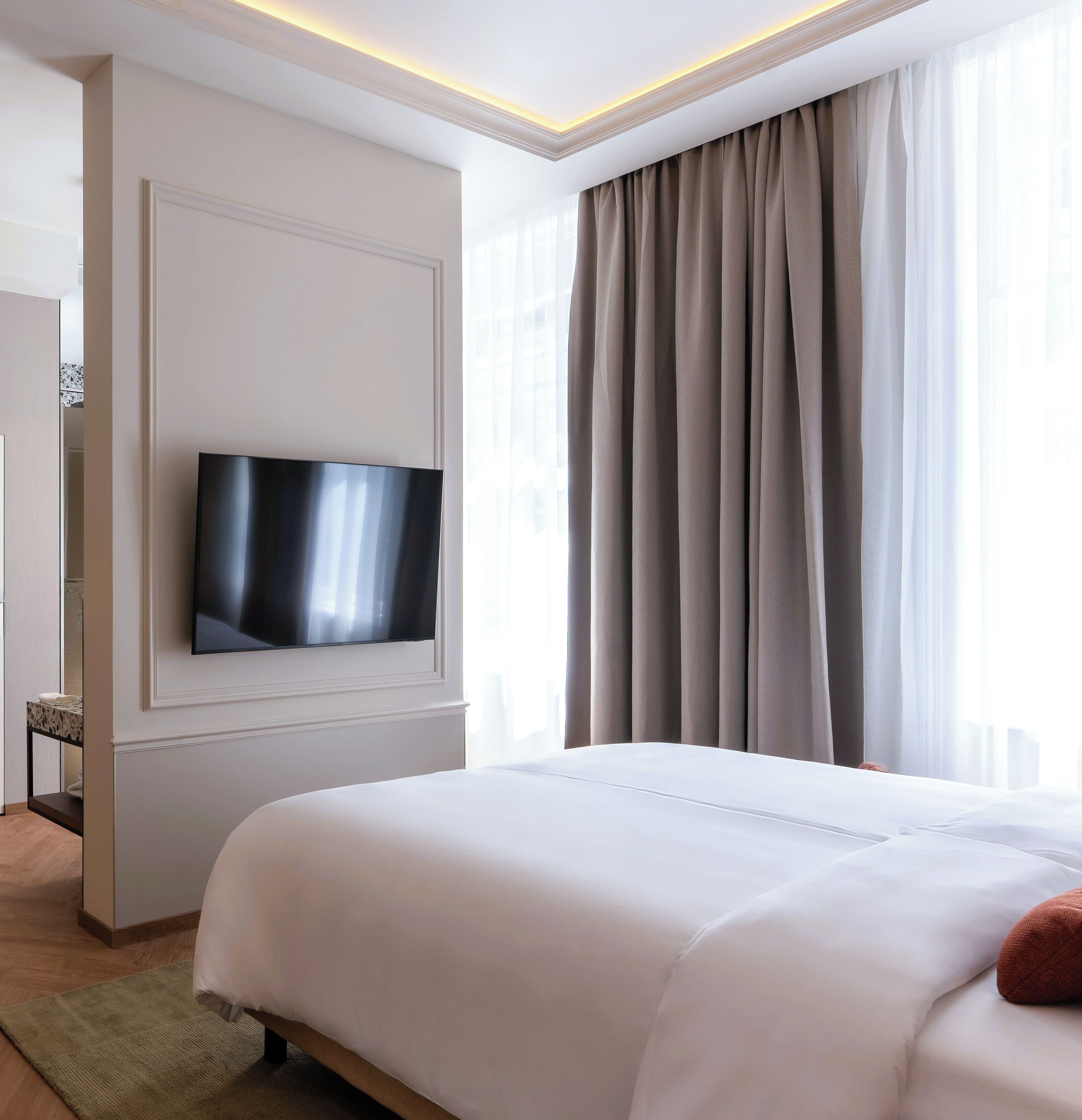 A modern hotel room with a flat-screen TV mounted on the wall, a large bed with white bedding, and a view of the city through sheer curtains.