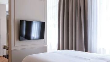 A modern hotel room with a flat-screen TV mounted on the wall, a large bed with white bedding, and a view of the city through sheer curtains.