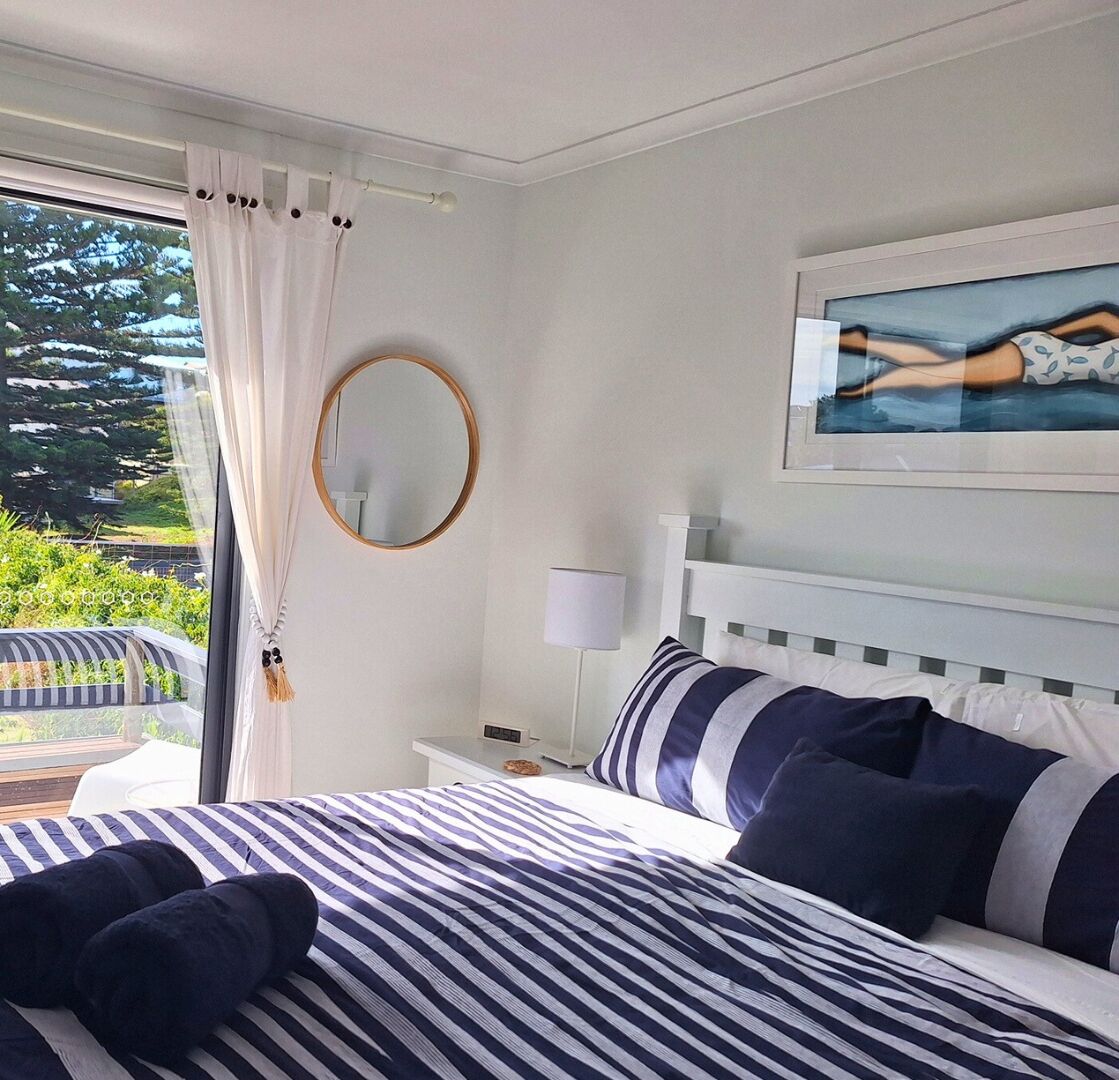 A hotel room with a bed, a bedside table, a lamp, a round mirror, and a painting on the wall.
