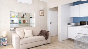 Una stanza con un divano color beige, uno scaffale in vetro con oggetti decorativi, un forno a microonde e una sedia trasparente in plastica.