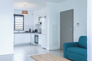 A modern kitchen with white cabinets, a built-in oven, and a microwave. There is a teal sofa in the living area. - Les Dunes - Front de Mer de Saint-Paul - Apartment sleeps 6 (Saint-Paul)