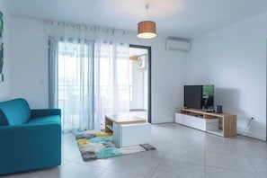 A modern living room with a teal sofa, a flat-screen TV on a wooden stand, and a large window with sheer curtains. - Les Dunes - Front de Mer de Saint-Paul - Apartment sleeps 6 (Saint-Paul)