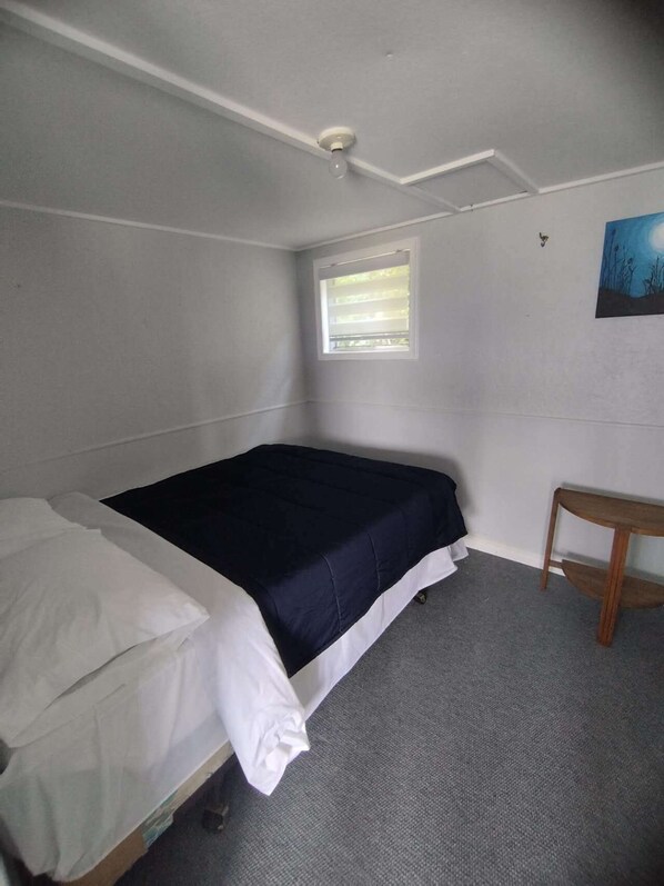 A small, single-bed room with a window, a bedside table, and a ceiling light fixture.