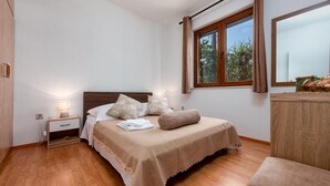 A neatly arranged bedroom with a wooden floor, a bed with a beige cover, a bedside table with a lamp, and a window with curtains.