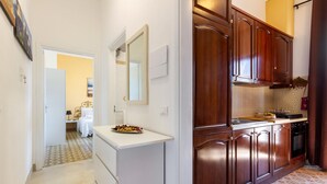 Un pasillo estrecho que conduce a un dormitorio con cama y mesita de noche. La habitación tiene una puerta blanca y un ventanal pequeño.