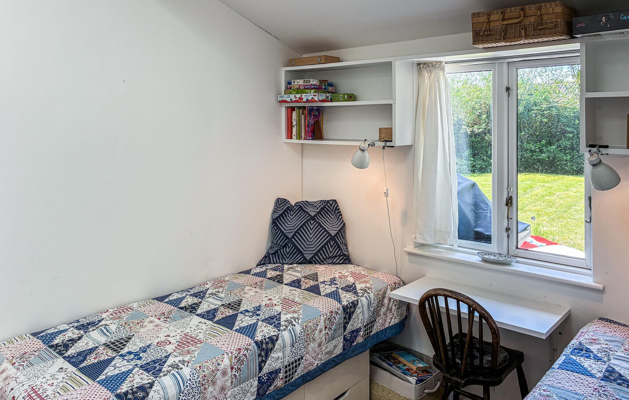 Une petite chambre bien rangée, avec un lit, un bureau et une chaise. Il y a une fenêtre avec des rideaux, une étagère remplie de livres et une lampe.