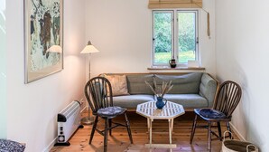 Een knus vertrek met een bank, twee stoelen, een kleine tafel, een radiator, een raam met uitzicht op groen en een gekaderd schilderij aan de muur.