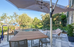 En udendørs terrasse med et træbord og stole, en stor parasol og et poolområde.