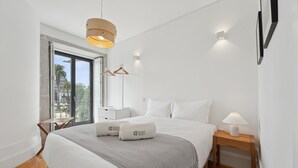 A modern bedroom with a large bed, a bedside table, a hanging light fixture, and a window with a view of palm trees.