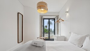 A modern hotel room with a large bed, a wooden-framed mirror, a hanging light fixture, and a view of palm trees through a sliding door.
