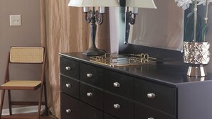 Una habitación con un aparador negro, un espejo, una lámpara, una silla y un jarrón con flores blancas.