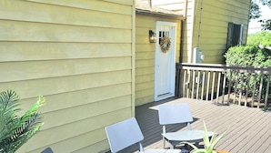 Una terraza con dos sillas grises y una mesa de madera, un tapete estampado y una casa con revestimiento amarillo y una puerta blanca.