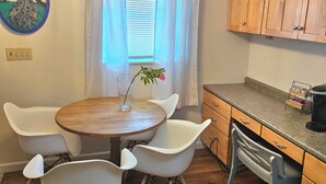 Un comedor con una mesa de madera y sillas blancas, una araña, una ventana con persianas y armarios de madera.