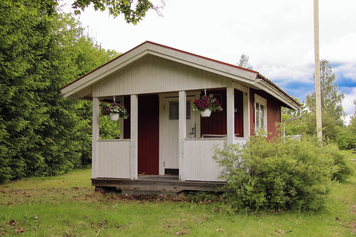 Eine kleine, einstöckige Holzhütte mit Veranda, umgeben von viel Grün.