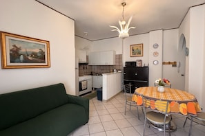 Un espace de vie compact avec un canapé vert, une table à manger recouverte d’une nappe orange et un coin cuisine doté de meubles blancs et d’une cuisinière.