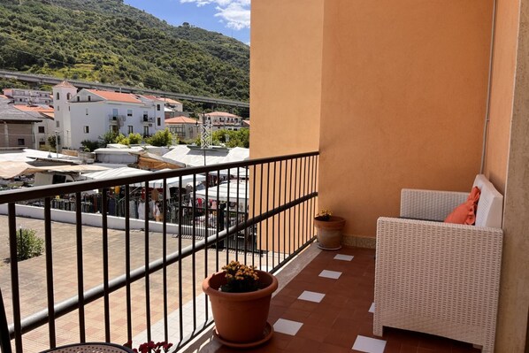 A balcony with a wicker sofa and potted plants, overlooking a street with buildings and a hill.