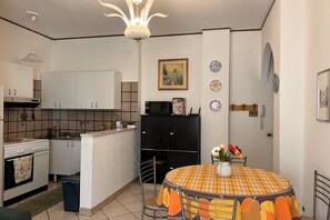 Une cuisine de taille modeste, avec des meubles blancs, un réfrigérateur noir et une table à manger recouverte d’une nappe à carreaux.