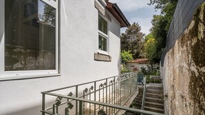 Un edificio blanco con una escalera de metal que lleva a una puerta, rodeado de follaje y un muro de piedra.