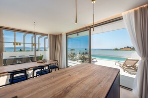 A modern dining area with a wooden table and chairs, large windows with a view of the sea, and a poolside lounge area. - Villa Mare Zadarvillas (Zadar)