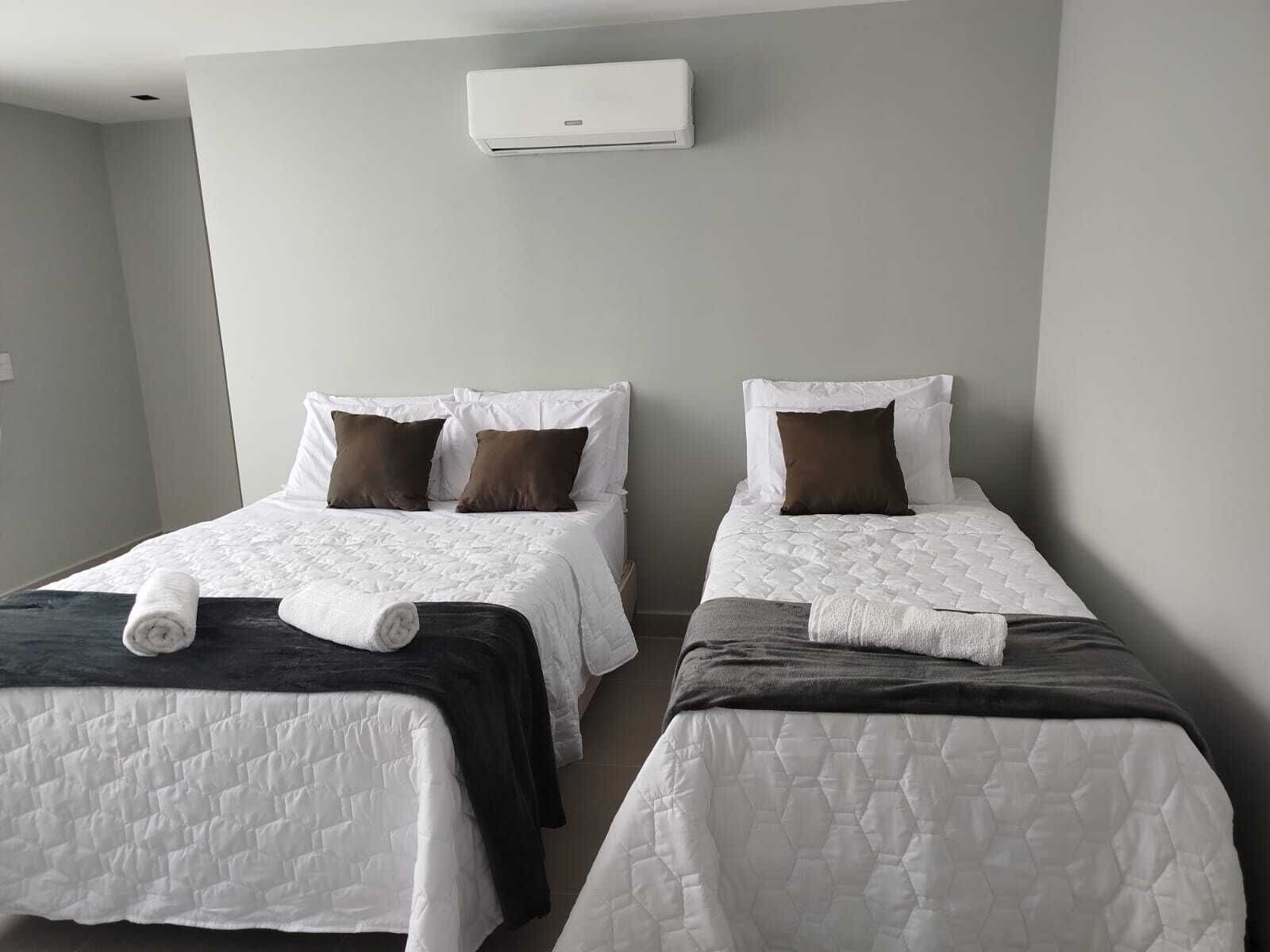Two single beds with white and dark bedding, a white air conditioner on the wall, and a plain grey wall.