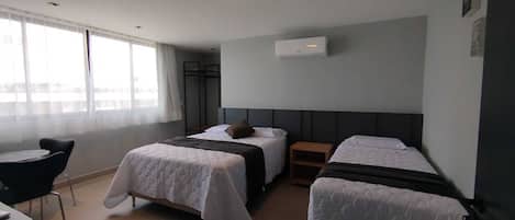 Habitación de hotel con dos camas, una mesita pequeña y una silla. Se ve una ventana con cortinas translúcidas, un equipo de aire acondicionado en la pared y un perchero.