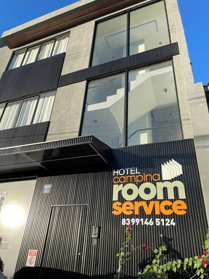 A modern hotel building with large windows, a prominent 'HOTEL campina room service' sign, and a contact number.