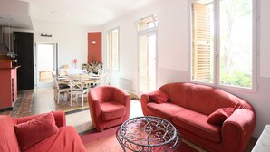 A spacious living area with a red sofa, a matching armchair, and a dining table with chairs. Large windows provide ample natural light. - House with character, spacious rooms, wooded garden, quiet, free parking (Argelès-sur-Mer)
