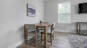 Un comedor con una mesa, sillas, una planta en maceta, una ventana con persianas y un televisor en la pared.