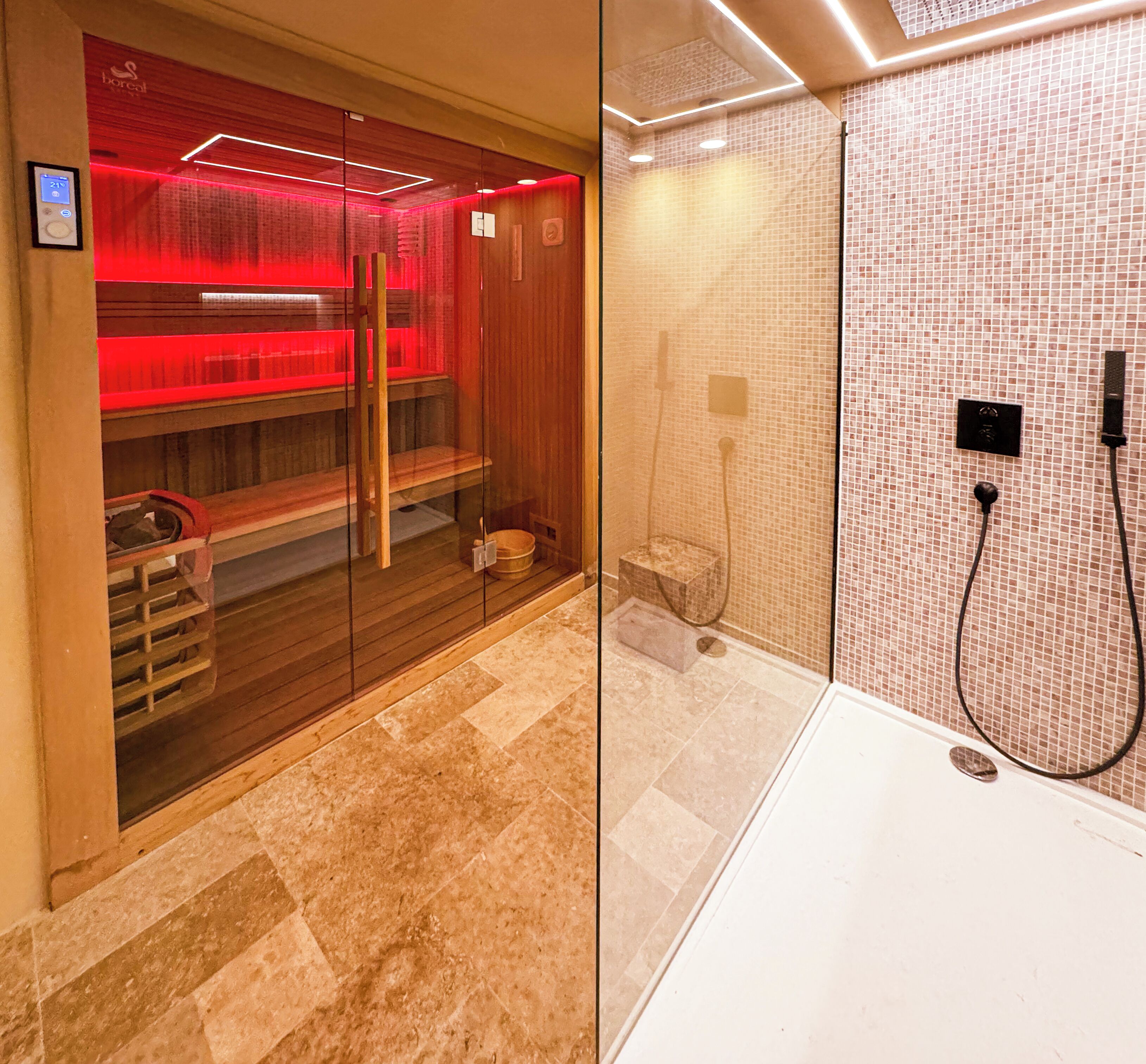 Una sauna con bancos de madera y un panel de vidrio, una bañera blanca con cabezal de ducha y paredes de azulejos.