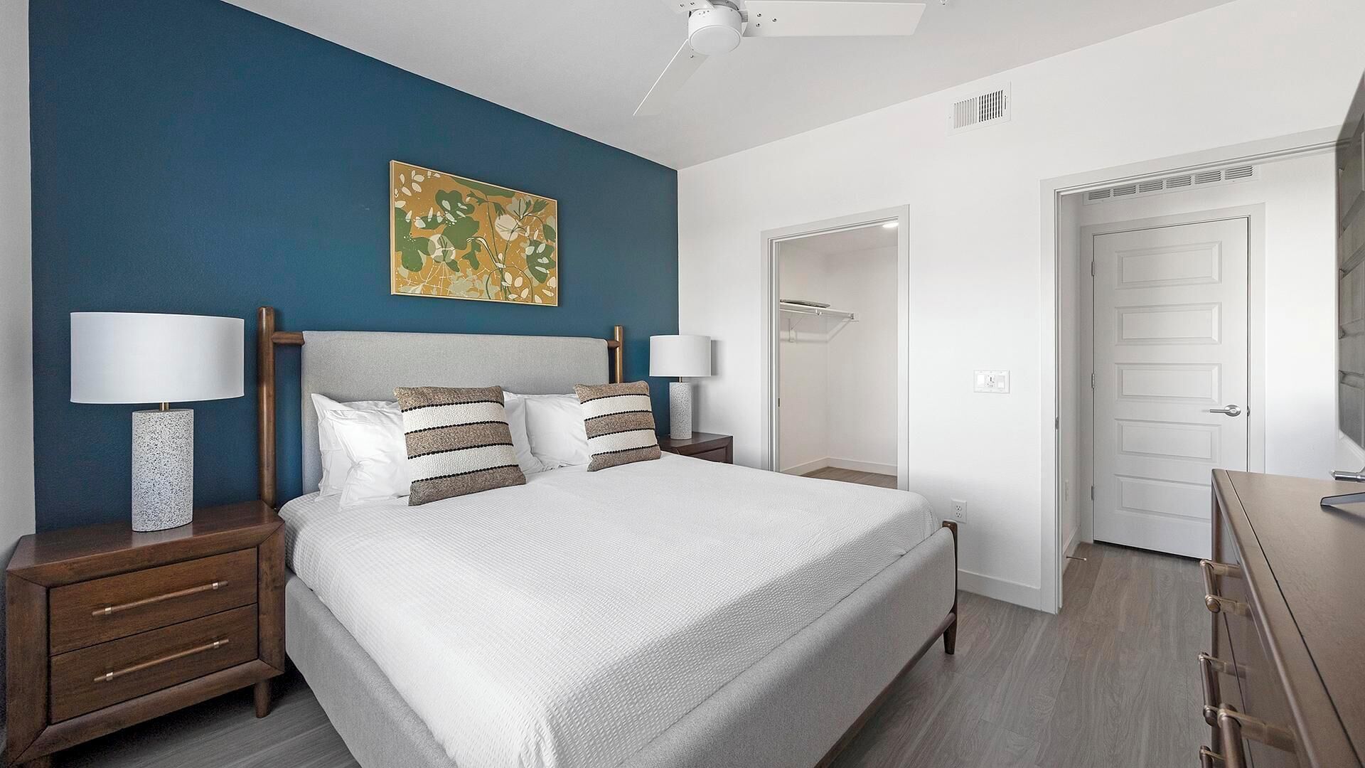 Une chambre à coucher comprenant un lit, deux tables de chevet, une commode, un ventilateur de plafond, une porte et un mur orné d’un tableau.